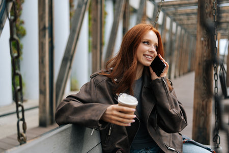 Stylish young redhead woman in leather jacket holding coffee cup and chatting on phone sitting on city swing at park on spring day. Happy red-haired female enjoy smartphone talking on park swing.の写真素材