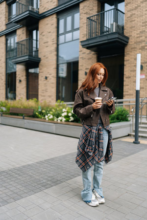 Full length vertical portrait of young woman with thoughtful expression standing in street, holding cup of coffee and using smartphone. Pensive redhead female waiting for someone outdoors.の写真素材