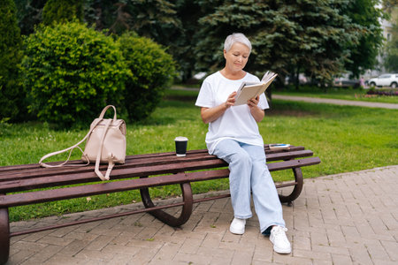 Full length portrait of gray-haired woman reading book in city park. Beautiful lady admiring green garden sitting on bench in summer day. Peaceful stylish pensioner holding literature resting outdoorsの写真素材