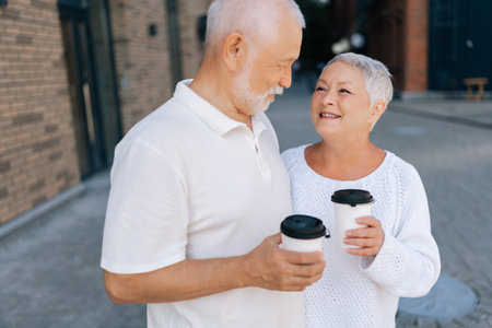 Handsome happy senior male with white beard enjoying coffee break with loved elderly wife in city street. Positive retired couple happily chatting and drinking coffee on urban street.の写真素材