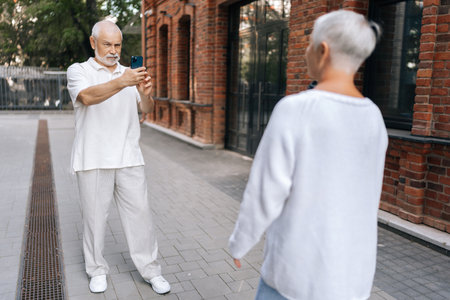 Elderly retired male photographer happily taking photo on smartphone of senior blonde woman holding coffee cup outside charming brick building, exploring urban landscapes, creating memories together.の写真素材