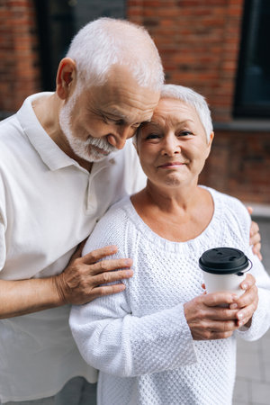 Vertical portrait of pretty elderly couple enjoying coffee on city street, standing hugging outdoors on warm sunny day, looking at camera. Concept of active aging and happy golden years.の写真素材