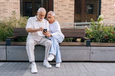 Portrait of cheerful elderly couple laughing sitting on city bench, sharing funny moment on smartphone, embodying senior love and togetherness through modern technology. Concept of retirement lifeの写真素材