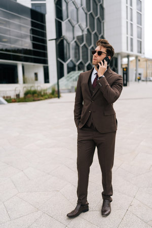 Full length vertical portrait of successful businessman in suit and sunglasses making call using mobile phone standing in city street on cloudy day. Concept of business communication and technology.の写真素材