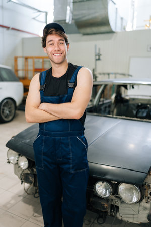 Vertical portrait of friendly young repairman in uniform and cap standing with crossed arms working in car service as mechanic, smiling looking at camera on blurred background of cars under repair.の写真素材