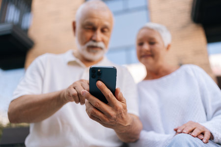 Low-angle view of cheerful senior married couple sitting at urban bench and using smartphone together on warm summer day, selective focus. Concept of modern technology, retirement life.の写真素材