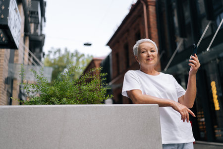 Relaxed senior woman using smartphone and smiling looking at camera while standing in street near flower bed, enjoying summer day in city environment, enjoying free time in retirementの写真素材