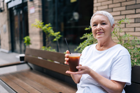 Portrait of serene senior woman enjoying iced coffee sitting on bench outside city cafe, enjoying moment of relaxation, smiling looking at camera on cozy summer day, enjoying free time in retirement.の写真素材