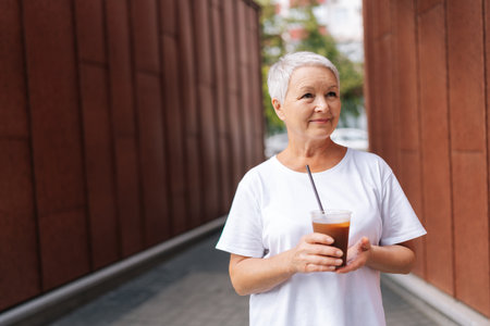 Portrait of stylish senior woman enjoying leisurely walk with iced coffee in city, radiating happiness and confidence on sunny day. Elegant elderly lady strolling with iced coffee in city alone.の写真素材