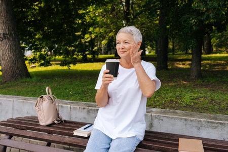 Portrait of happy smiling 60s female sitting on bench and drinking coffee outdoor in park on summertime. Positive attractive older woman drinking takeaway coffee on wooden bench in park.の写真素材