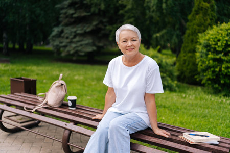 Thoughtful elderly woman with silver hair relaxing on park bench with backpack, books, and cup of coffee, savoring tranquility. Serene European gray-haired older female enjoying moment of relaxation.の写真素材