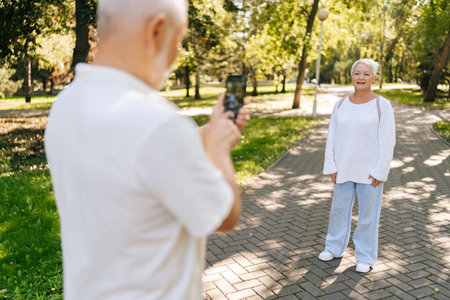 Beautiful elderly couple happily taking photos in park on sunny summer day, creating memories with mobile phone, exuding love and companionship. Active senior people enjoying retirement together.の写真素材