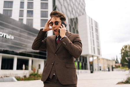 Angry young businessman in suit and sunglasses experiencing extreme stress while talking on mobile phone, pulling hair and yelling outside of office building. Concept of business communication.の写真素材