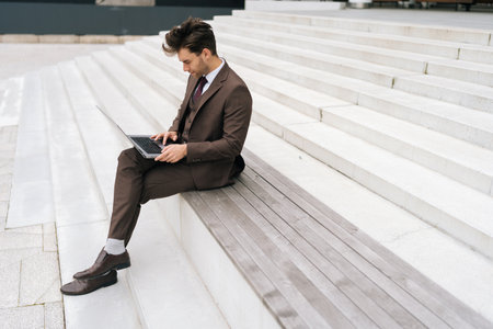 Wide shot of focused businessman working remotely while sitting on steps in urban setting, typing on laptop computer, focused looking to screen. Concept of business communication and technology.の写真素材