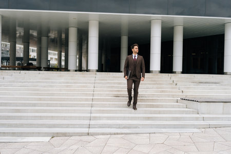 Portrait of confident successful businessman in brown suit walking down stairs of modern office building holding laptop in hand. Business man descending stairs of contemporary office building.の写真素材