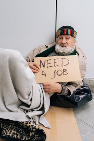 Vertical portrait of despair elderly homeless man with sign seeking employment on urban street, appearing distressed and hopeless amid economic downturn. Concept of alone poor unhappy retirement.の写真素材
