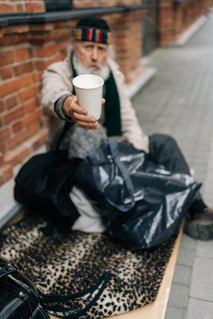 Full length vertical portrait of older homeless man sitting on urban street with belongings. Sad aged tramp seeking assistance, holding disposable cup and begging for alms. Concept of poor retirement.の写真素材