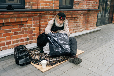 Wide shot of homeless senior male sitting alone at cold autumn day on city street, holding legs for warmth, expression showing despair and loneliness, emphasizing challenges of urban poverty.の写真素材