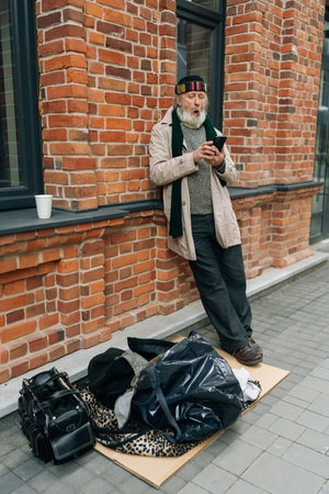 Full length vertical portrait of senior homeless man with white beard using mobile phone looking to screen while leaning on brick wall in city street, with belongings lying on cardboard on ground.の写真素材