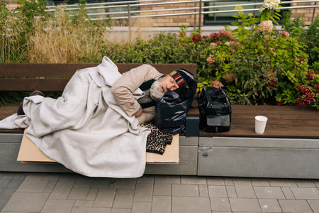 Exhausted older homeless man peacefully sleeping on weathered bench outside in urban city streets on cold day. Tired aged beggar male lying outdoors. Concept of raw reality of poverty and despair.の写真素材