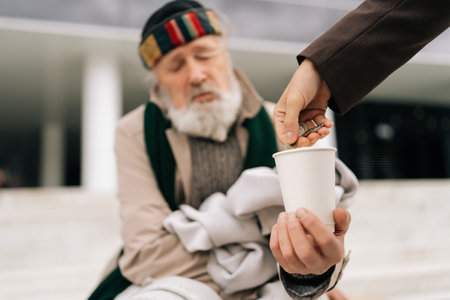 Cropped shot of unrecognizable businessman in suit giving alms to homeless senior man wearing dirty clothes holding paper cup, sitting on city street. Business man generously donates to beggar male.の写真素材