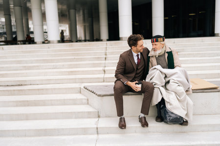 Kindness smiling businessman listening and smiling as talking with elderly homeless male, demonstrating empathy and kindness in heartwarming moment of human interaction, sitting on urban steps.の写真素材