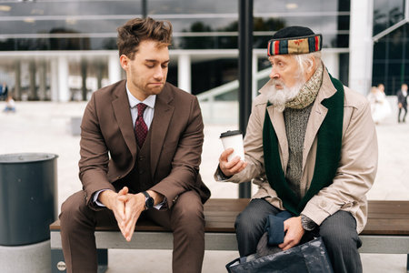 Handsome businessman in formal attire attentively engages in conversation with senior homeless man holding coffee cup, while seated on bench in public area, on cloudy day.の写真素材