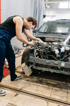 Vertica rear view of focused mechanic male in blue uniform inspecting car engine in workshop,...