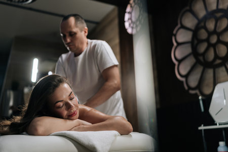 Beautiful young woman relaxing with hand massage at beauty spa lying with closed eyes. Professional masseur giving back massage to female client experiencing therapeutic wellness in beauty parlor.の写真素材
