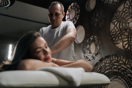 Serene beautiful young woman relaxing during back massage in spa with soft light, looking at camera. Charming brunette female getting relaxing massage. Concept of rest with spa treatment.の写真素材