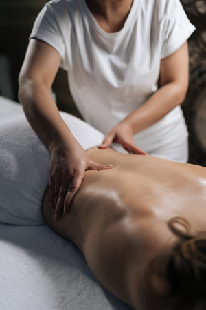 Vertical cropped shot of unrecognizable masseur spreading massage oil, gently working on woman client back during soothing spa treatment in serene, softly illuminated wellness environment.の写真素材