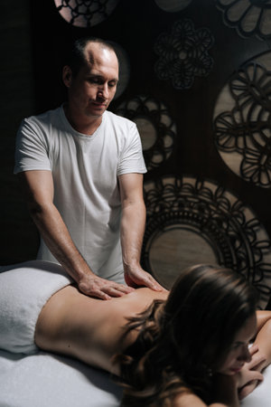 Professional masseur giving back massage to female client lying on massage table in modern spa center with dark interior. Serene young woman enjoying relaxing treatment at luxury beauty parlor.の写真素材