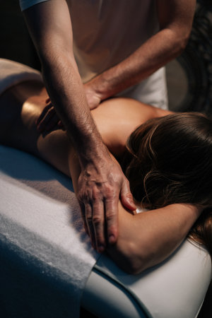 Vertical cropped shot of massage therapist performing deep tissue technique on female shoulder,...