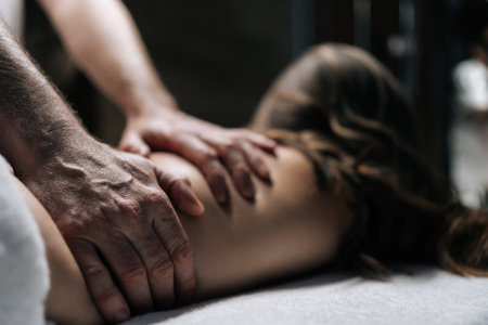 Masseur male performing deep tissue technique on female back, targeting muscle tension and promoting wellness during bodywork session in spa with dark interior. Concept of luxury professional massage.の写真素材