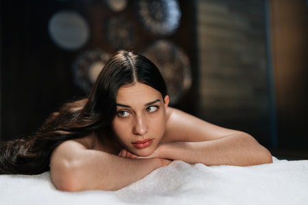 Closeup face of calm young woman lying on massage table with hands under chin preparing for relax massage procedure in spa salon. Charming brunette female awaiting beauty treatment in wellness center.の写真素材