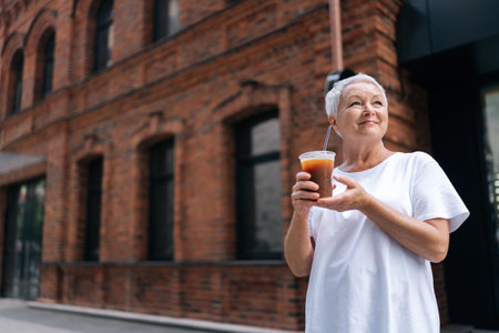 Low angle view of happy senior woman holding cup with iced coffee and looking up with smile on face, enjoying moment of relaxation outside on background of residential building on summer day.の写真素材