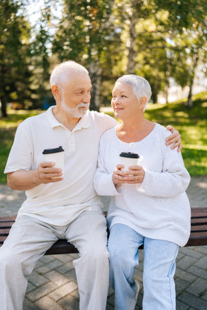 Vertical portrait of happy gray-haired couple enjoying sunny day in park, sitting on bench with coffee, surrounded by green trees, embracing and smiling, embodying love and happiness in retirement.の写真素材