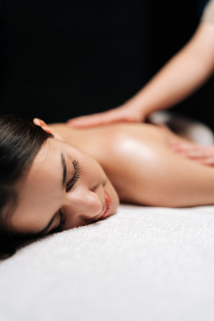 Closeup vertical shot of carefree woman client relaxing during back massage in spa salon with dark interior. Professional massage therapist providing therapeutic back massage to relaxed female.の写真素材