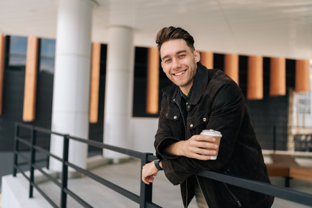 Portrait of happy young man holding takeaway coffee cup leaning on metal railing standing by modern building, smiling looking at camera. Serene dreaming male drinking enjoying coffee outdoors.の写真素材