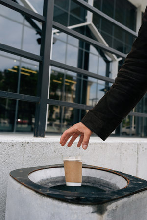 Close-up vertical cropped shot of unrecognizable busines man throwing disposable coffee cup into street trash can near office building, demonstrating responsible waste disposal and urban cleanliness.の写真素材