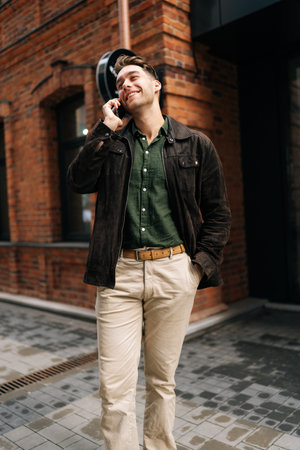 Vertical portrait of happy businessman walking down street while talking on mobile phone, enjoying conversation on a sunny day in urban environment. Concept of modern successful urban lifestyle.の写真素材