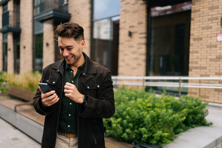 Portrait of cheerful young man standing on city street, with smile on face using smartphone to check messages, browse internet. Concept of modern communication and urban lifestyle.の写真素材