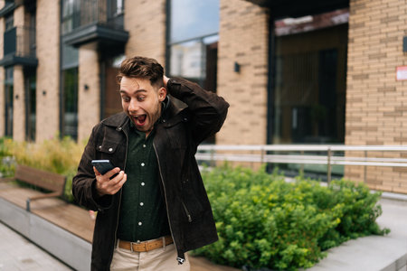 Happy excited young man standing in city street, holding mobile phone and reacting with astonishment to some unexpected news just received, looking to smartphone screen with amazed expression.の写真素材