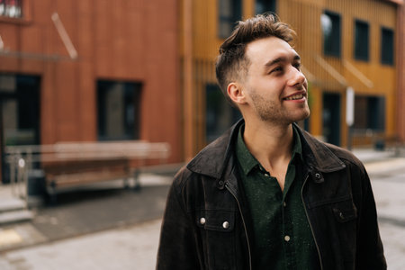 Portrait of handsome bearded young man wearing dark jacket and green shirt smiling looking away, standing in urban setting filled with contemporary architecture and cheerful atmosphere.の写真素材
