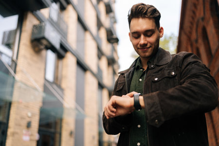 Low-angle portrait of fashionable young man in brown leather jacket checking smartwatch outside modern building, possibly during workday commute or break. Concept of modern successful city lifestyle.の写真素材