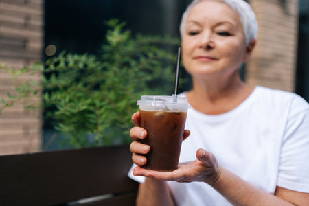 Selective focus portrait of senior woman drinking refreshing iced coffee sitting at outdoors cafe,...