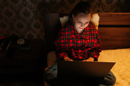 Top view of preteen girl in checkered pajamas smiling looking to screen while using laptop sitting on bed in dark bedroom at night, face illuminated by screen light, creating cozy atmosphere.の写真素材