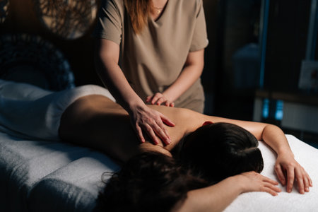 Closeup of unrecognizable masseuse applying massage oil back of young woman lying on massage table, providing relaxing and therapeutic body treatment in spa salon with dark interior.の写真素材