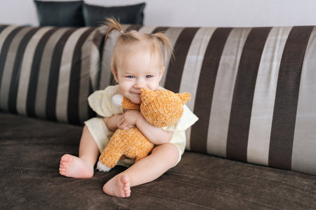 Portrait of adorable baby girl hugging handmade knitted teddy bear toy while sitting on comfortable sofa at home, enjoying quiet moment of play and affection. Concept of simple pleasures childhood.の写真素材