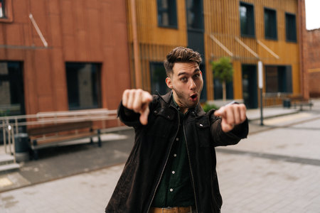Portrait of cheerful young man pointing fingers at camera with funny expression standing in city setting, creating candid and engaging shot. Handsome happy male gesturing outside looking at camera.の写真素材